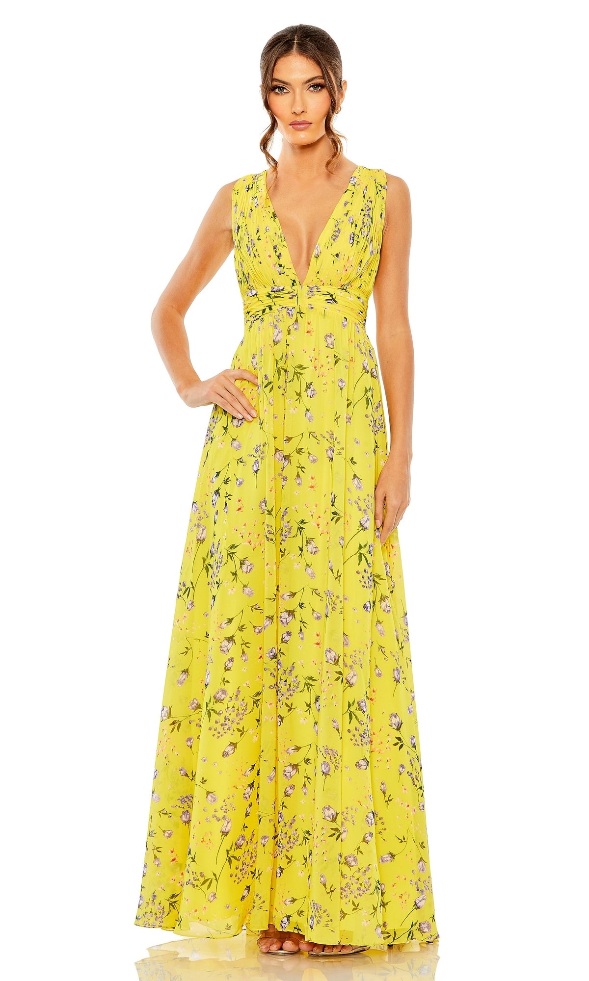 A woman stands confidently with one hand on her hip, wearing the Long Formal Dress 56011 by Mac Duggal—a sleeveless, floor-length yellow floral dress featuring a deep V-neckline and fitted A-line silhouette.